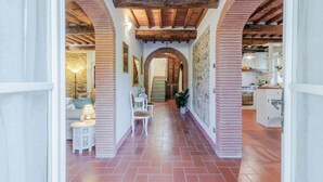 A corridor with a tiled floor, brick walls, and wooden beams on the ceiling. There are framed pictures on the walls and a small table with a lamp. - Villa Dondolino in Lucca (Lucca)