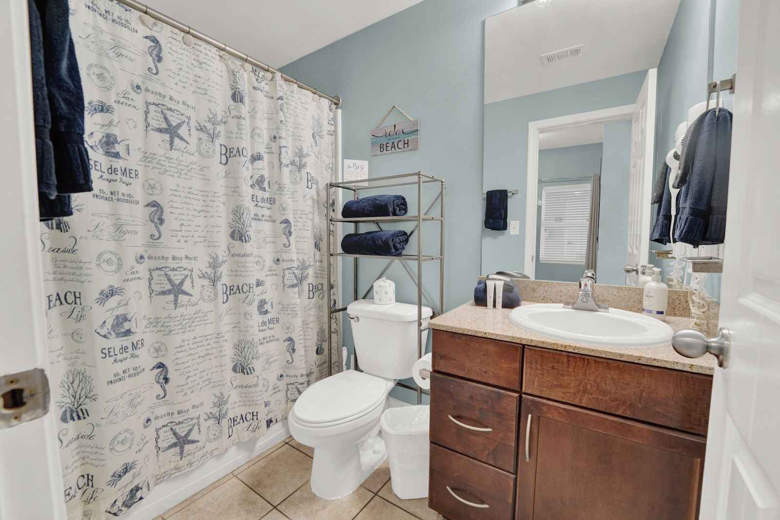 Une salle de bain équipée d’un rideau de douche orné de motifs représentant la plage et la vie marine, de toilettes, d’un lavabo avec un miroir et d’un porte-serviettes.