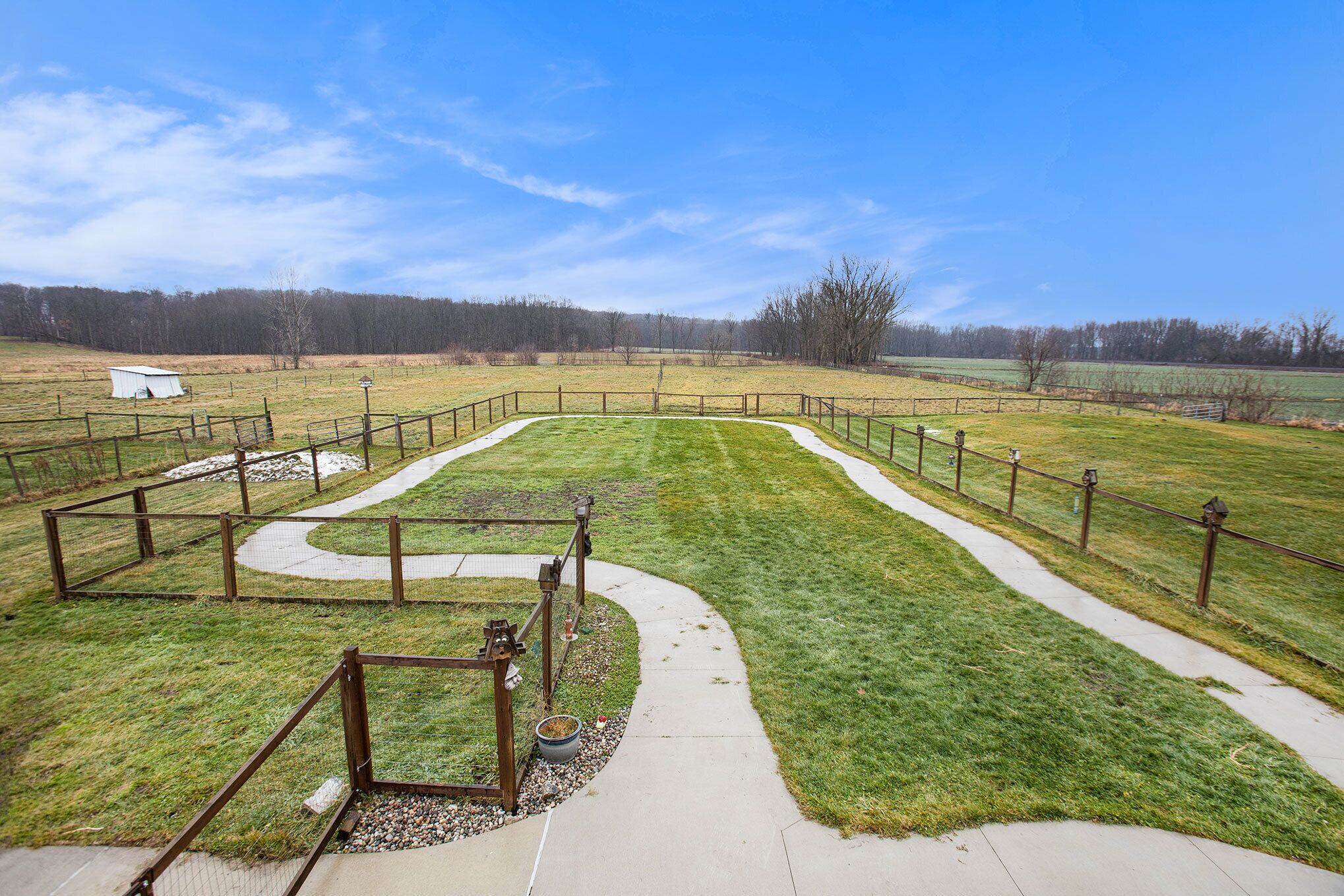 A fenced outdoor area with a paved walkway, a small structure, and a grassy field.