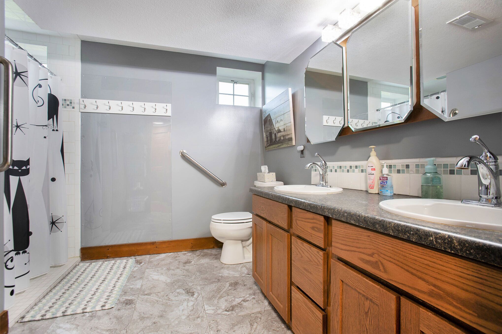 A bathroom with a white toilet, wooden cabinets, a granite countertop, a mirror, and a shower curtain with a cat design.