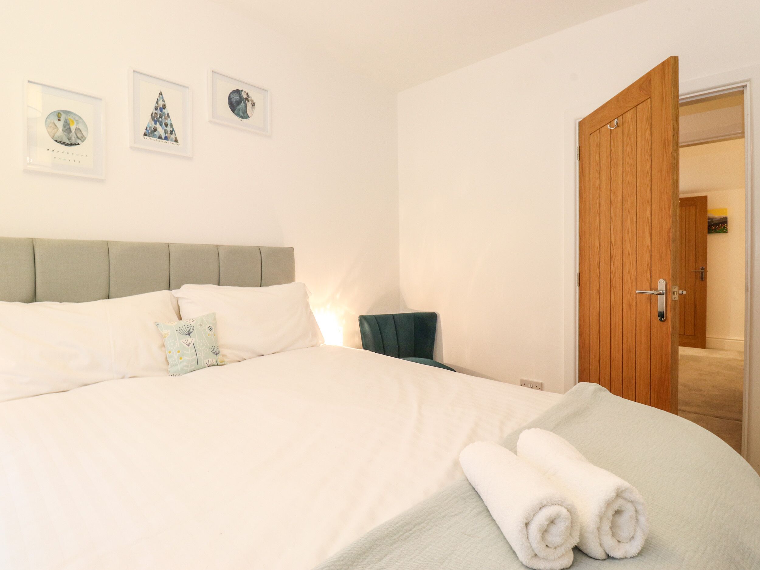 Una cama bien hecha con sábanas blancas y dos toallas enrolladas, una puerta de madera y tres cuadros en la pared.