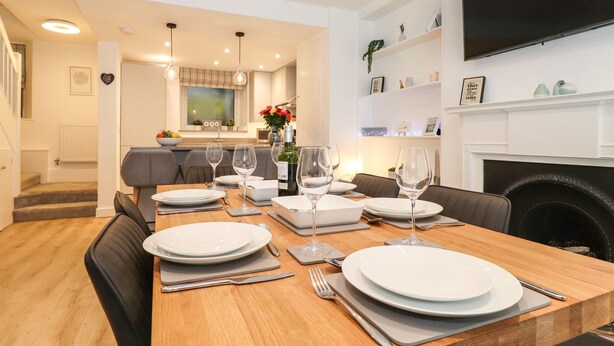 A dining area with a wooden table set for four, a fireplace, and a kitchen in the background.