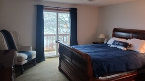 A bedroom with a wooden bed, a chair, a nightstand with a lamp, and a view through a window with curtains. - Riverfront house  with  kyacks  fire pit and gym (Wareham)