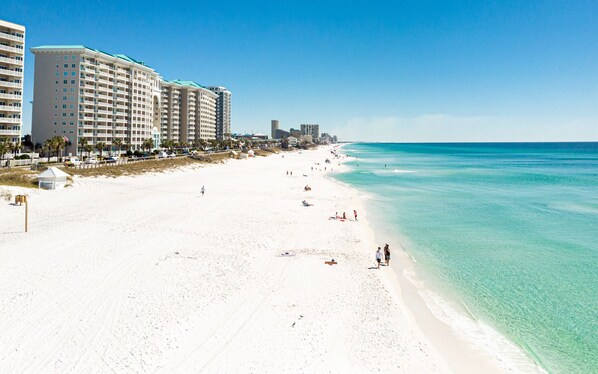 Una playa con arena blanca y agua turquesa cristalina. Varias edificaciones de varios pisos con balcones bordean la costa.