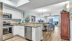 Cocina moderna con gabinetes blancos, encimera de granito y electrodomésticos de acero inoxidable. Cuenta con un comedor con vista a la playa.