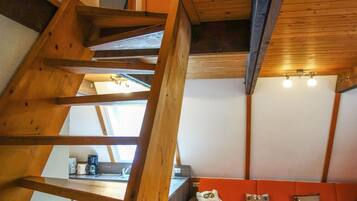 Un escalier en bois mène à une chambre aménagée en mezzanine, dotée d’une lucarne, d’un coin cuisine et d’un canapé rouge.