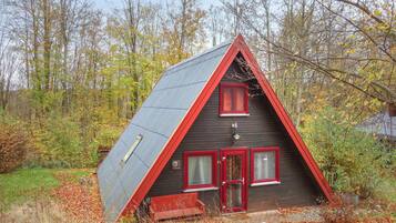 Une petite cabane triangulaire en bois, avec un toit rouge et deux fenêtres à cadre rouge, entourée du feuillage automnal.
