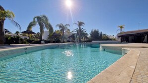 A clear swimming pool with a tiled edge, surrounded by palm trees and a building with a tiled roof. - S o Rafael Beach House (Albufeira)