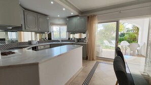 A modern kitchen with a central island, large windows, and a sliding glass door leading to an outdoor seating area. - S o Rafael Beach House (Albufeira)