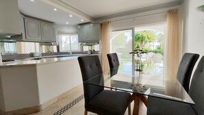 A modern kitchen with white cabinets, a dining area with glass tables and chairs, and a view of a garden through large windows. - S o Rafael Beach House (Albufeira)