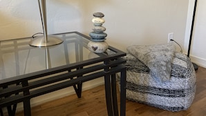 A glass-top table with a metal lamp, a stack of stones with inscriptions, and a woven pouf against a plain wall. - The Grand Adventure (Grand Junction)