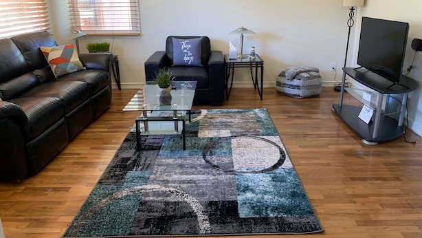 A living room with a black leather sofa, a black armchair, a glass coffee table with a plant, a colorful rug, a painting on the wall, and a TV stand. - The Grand Adventure (Grand Junction)