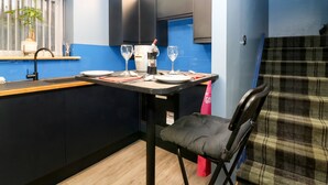 A compact kitchen with a dining table set for two, a coffee maker, and a window with a plant. - The Cavern at the Tavern (Blairgowrie)