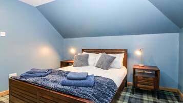 Quarto com cama de madeira, paredes azuis, tapete xadrez e duas mesas de cabeceira com abajures.