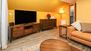 A living room with a flat-screen TV, wooden entertainment unit, orange sofa, and a small table with a lamp. - The Cavern at the Tavern (Blairgowrie)