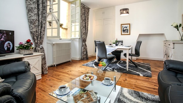 Una sala moderna con una mesa de centro de vidrio, sofás de cuero, un comedor con una mesa y sillas blancas, un radiador y una ventana con cortinas.