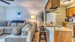 Een moderne woonkamer met een bank, een salontafel en een vloerlamp. Er is een keukenhoek met houten kastjes en een magnetron.