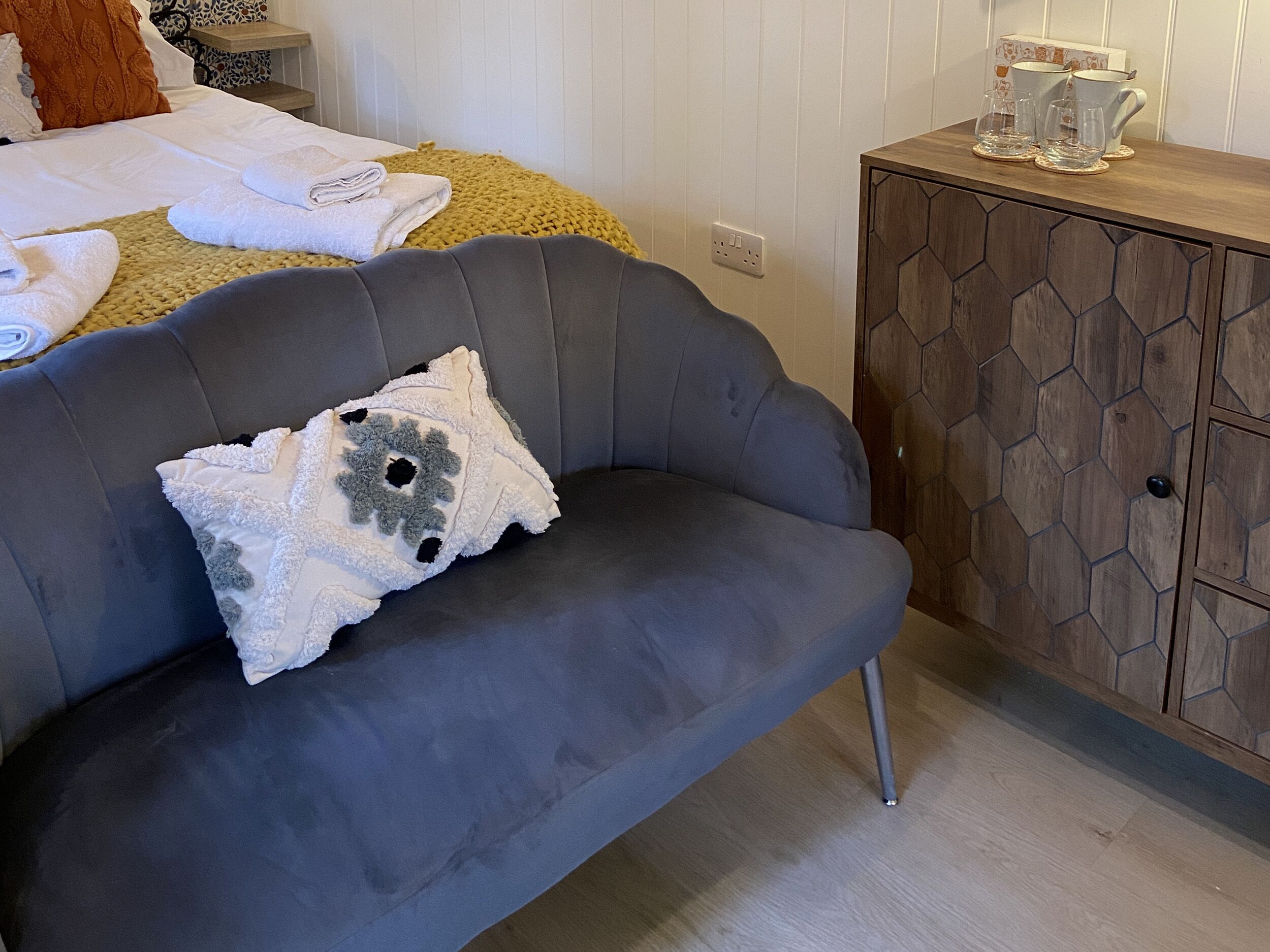 Un sofá gris con un cojín decorativo, un aparador de madera con diseños hexagonales y una cama con toallas enrolladas y una manta amarilla.