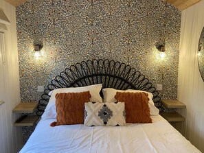 Ein Bett mit weißer Bettwäsche, zwei braune Kissen und ein Zierkissen mit geometrischem Muster. Der Raum zeichnet sich durch eine Wand mit Musterung und ein schwarzes Metall-Kopfteil aus.
