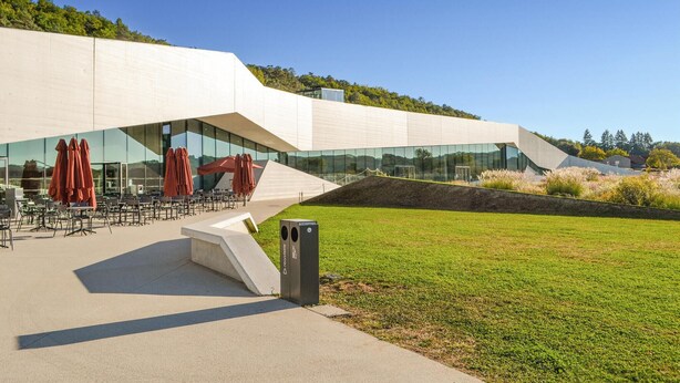 Un bâtiment moderne doté d’un toit en pente, d’un espace extérieur aménagé avec des parasols rouges et d’un paysage verdoyant.