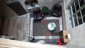 Une petite pièce aux murs de pierre, dotée d’un sol carrelé, d’une table ronde sur laquelle sont posés des magazines, de deux fauteuils en cuir, d’une petite plante et d’une fenêtre grillagée.