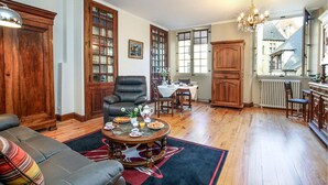 Un salon chaleureux avec un canapé en cuir, une table à manger dressée pour le petit-déjeuner, des meubles en bois et une grande fenêtre donnant sur un bâtiment à l’extérieur.