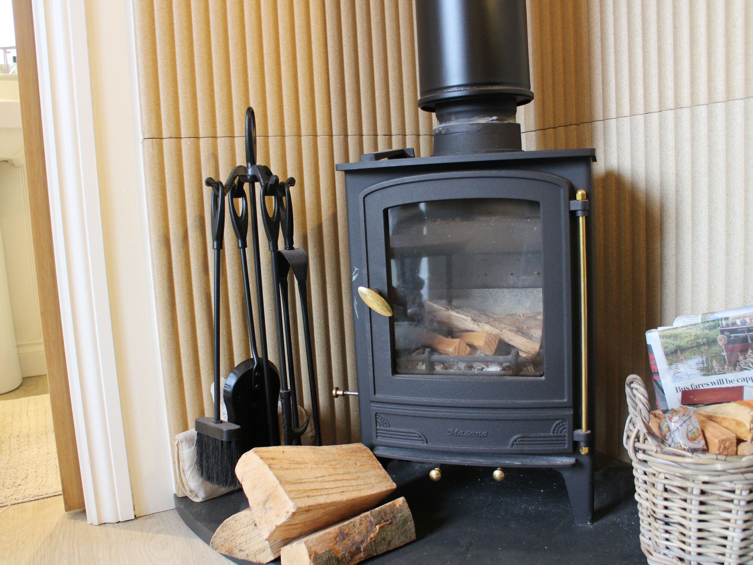 Una estufa de leña con troncos, una cesta de leña y un juego de herramientas para chimenea.
