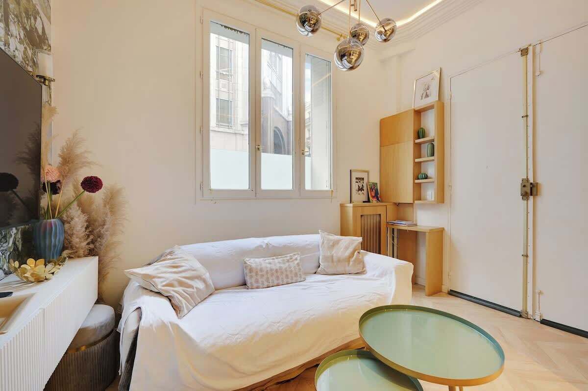 Une pièce avec un canapé blanc, une table en verre, un meuble en bois et une fenêtre donnant sur des immeubles.