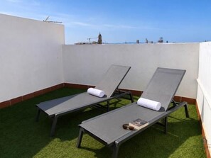 Two sun loungers with towels on a rooftop terrace with artificial grass and a clear sky.