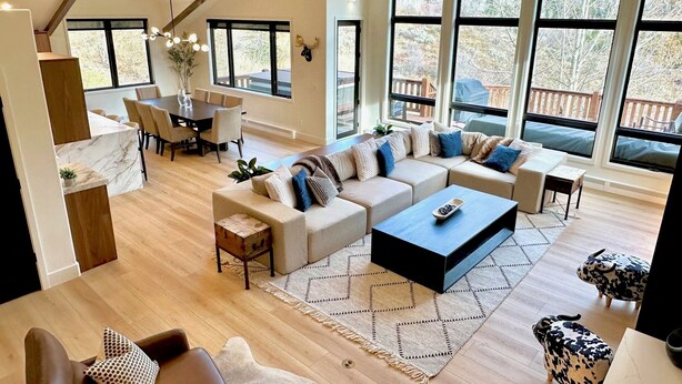 A spacious living area with a large sofa set, a dining table, and a coffee table. There are large windows providing natural light and views of the outdoors. - Luxe Vail! 5 minutes to town. Dual Primary - Hot Tub - Free Ski Bus! Sleeps 10 (Vail)
