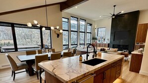 A modern kitchen with a marble island, wooden cabinets, and large windows offering a view of the outdoors. - Luxe Vail! 5 minutes to town. Dual Primary - Hot Tub - Free Ski Bus! Sleeps 10 (Vail)