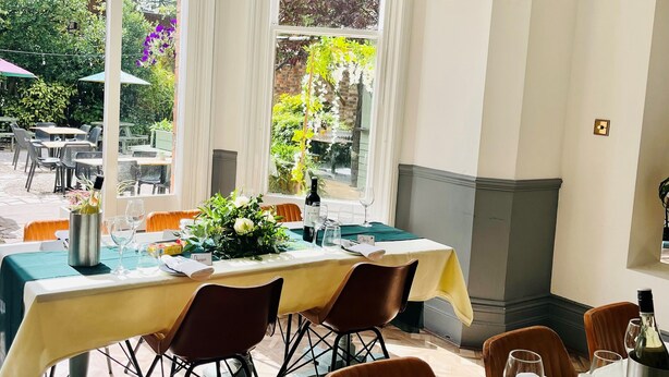 A dining area with a table set for two, a chandelier, and a view of the garden through large windows. - Blue Piano (Birmingham)