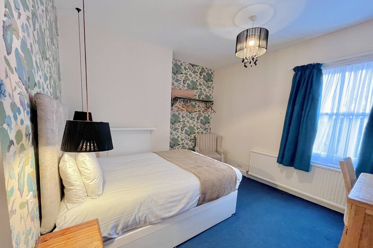 A bedroom with a bed, a hanging lamp, a chair, a radiator, and a window with curtains.