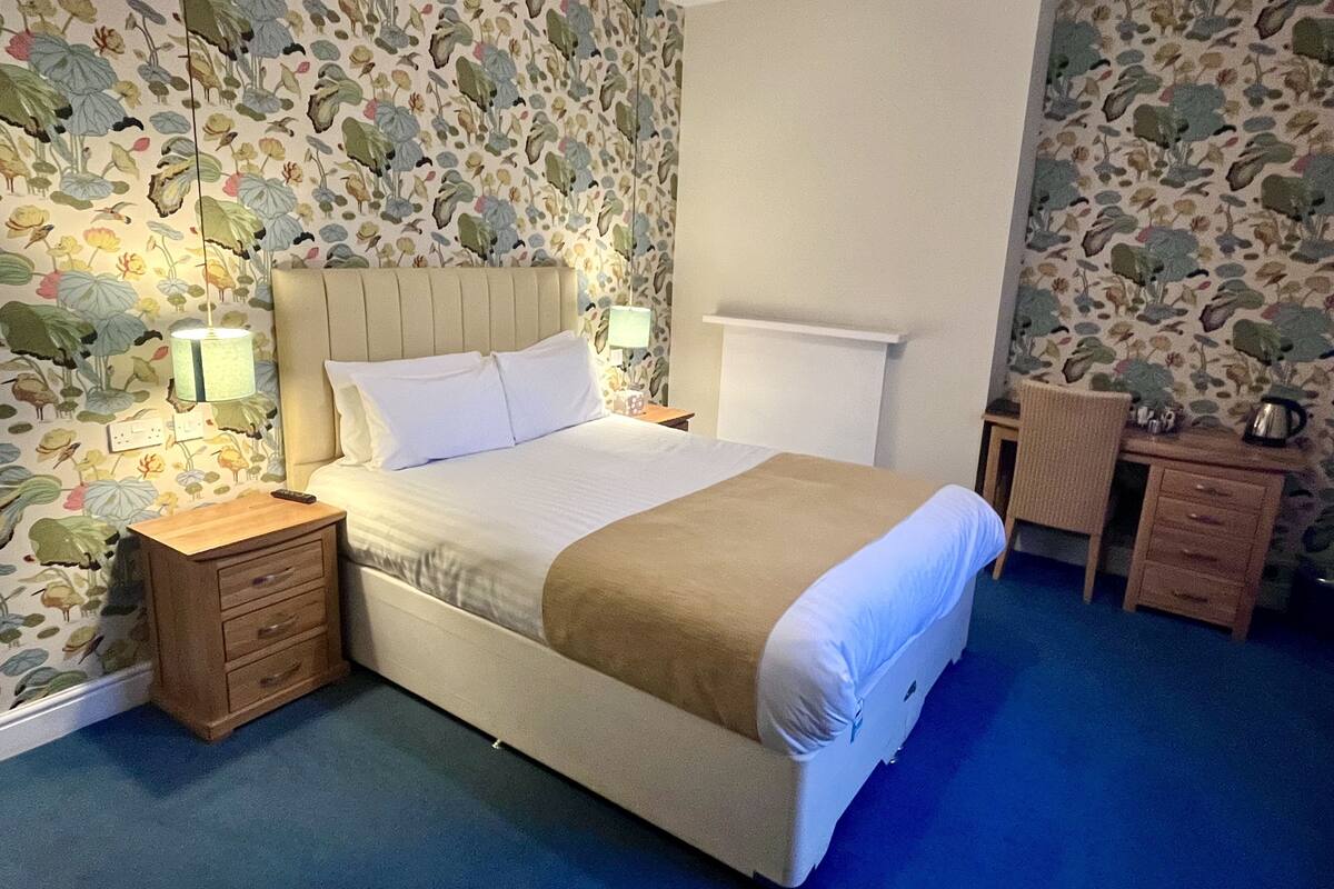 A neatly made bed with white linens and a beige throw blanket, a bedside table with a lamp, and a small desk with a chair. The room has floral wallpaper and a blue carpet.