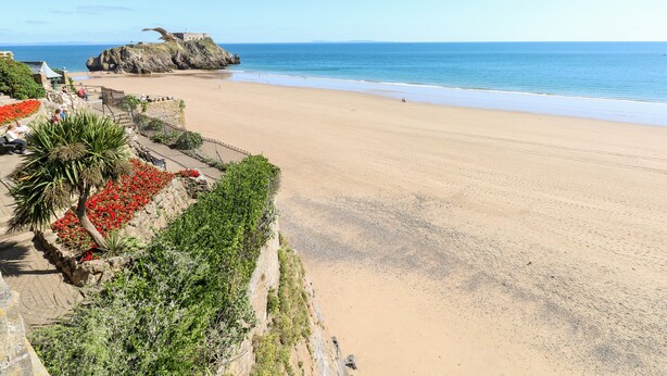 A beach with a clear view of the ocean, a rocky outcrop, and a pathway with red flowers. - Rosemary Cottage (Tenby)