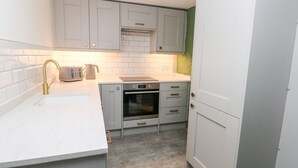 A compact kitchen with white cabinets, a stainless steel oven, a toaster, and a sink with a gold faucet. - Rosemary Cottage (Tenby)