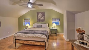Une chambre à coucher avec un lit, un ventilateur de plafond, deux tables de chevet avec des lampes, une fenêtre avec des rideaux et un vase rempli de fleurs placé sur une table d’appoint.