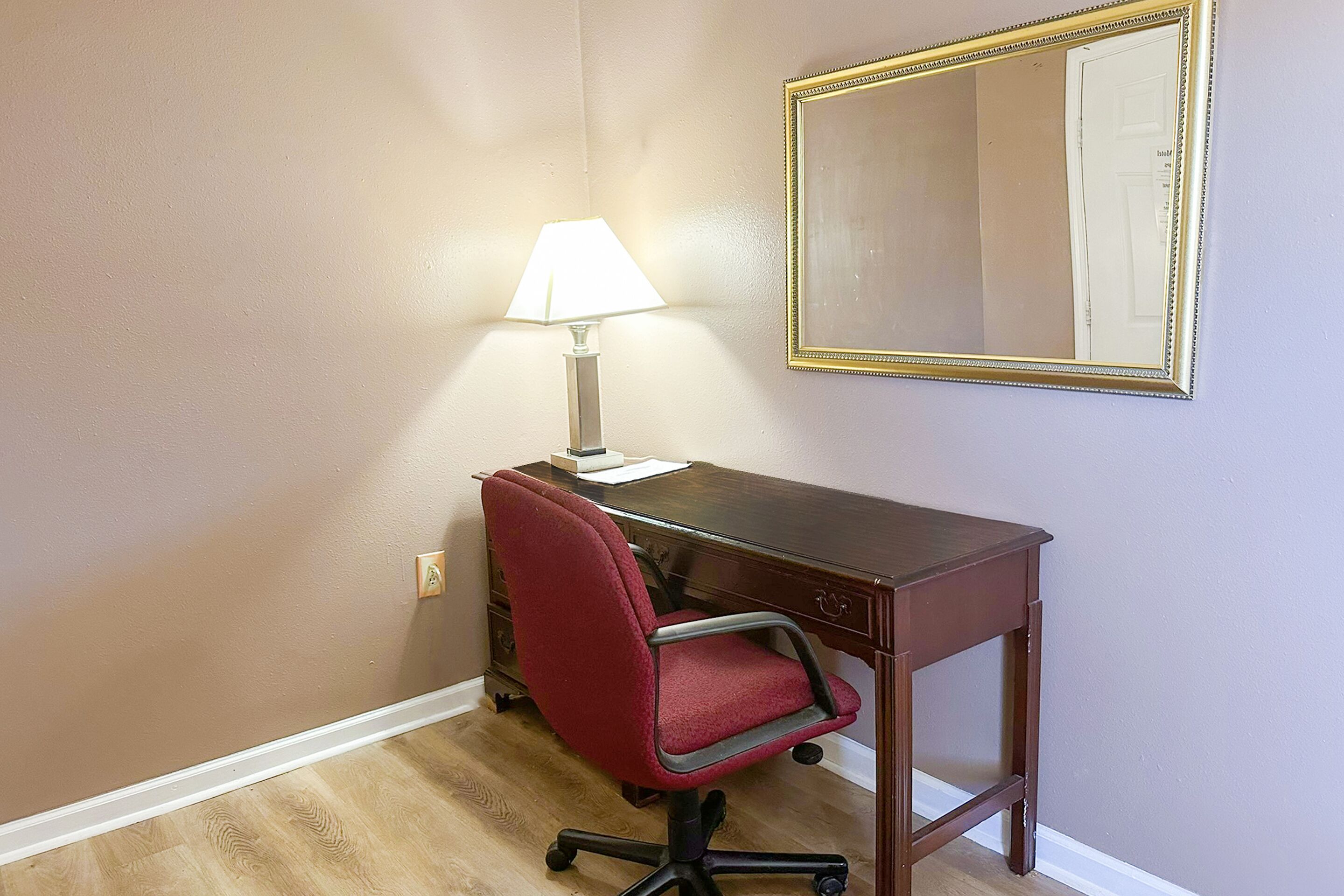 A room with a wooden desk, a red office chair, a lamp, and a mirror on the wall.