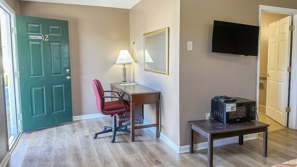 Una habitación con puerta verde, un escritorio con lámpara, una silla, un televisor montado en la pared y un microondas sobre una mesita.