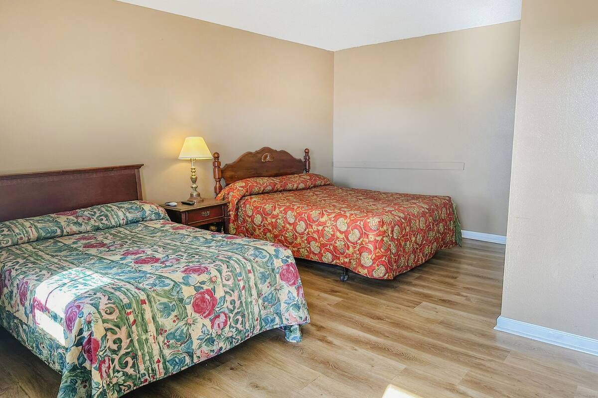 A room with two beds, a nightstand with a lamp, and wooden flooring.
