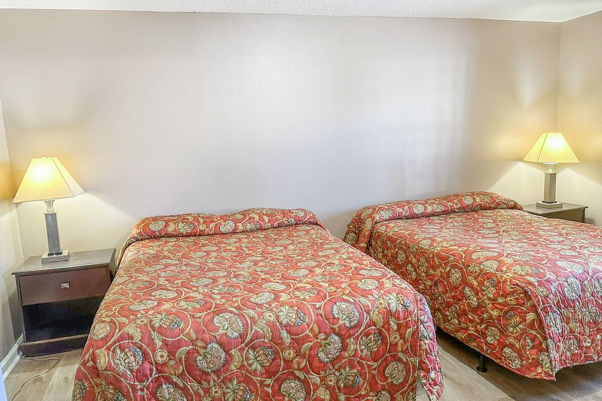 A room with two beds, each with a patterned bedspread, two bedside tables with lamps, and a plain wall.