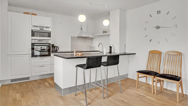 A modern kitchen with white cabinetry, a central island with bar stools, a built-in oven, and a wall clock. - 3 bedroom beautiful home in Bryrup (Bryrup)
