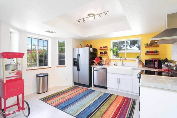 Eine moderne Küche mit weißen Schränken, einem Edelstahl-Kühlschrank, einer Spülmaschine und einem roten Küchenmixer. Fenster bieten Blick auf Grünflächen.