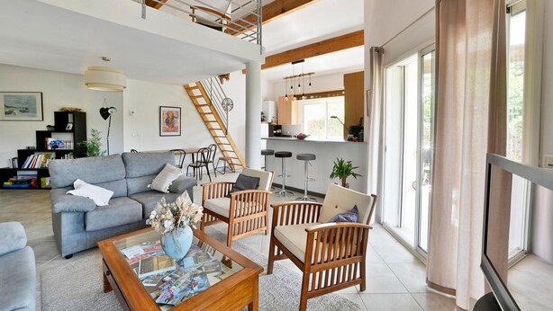 Un vaste espace de vie comprenant un canapé gris, des chaises en bois, une table basse avec des magazines, un escalier et un coin cuisine avec des tabourets de bar.
