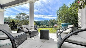 Une terrasse couverte, aménagée avec des meubles en osier, une petite table et offrant une vue sur un jardin et des arbres.