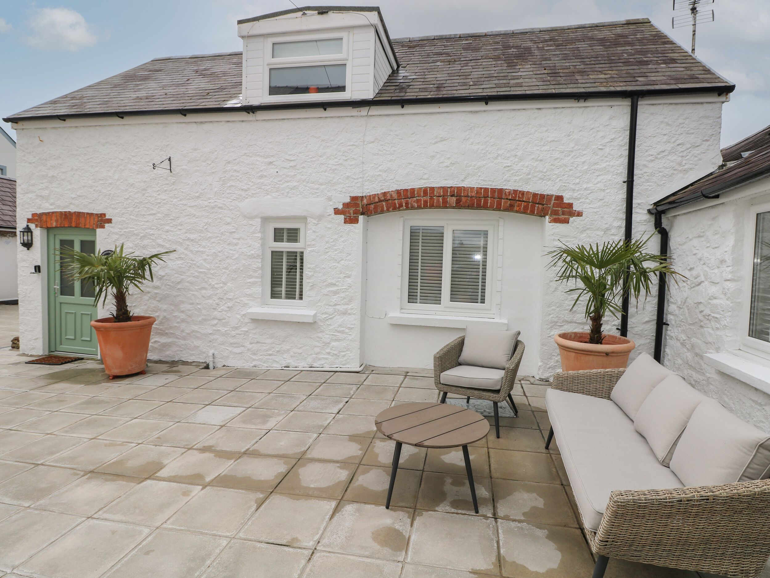 A white house with a tiled patio, two potted plants, a wicker sofa, and a small table.