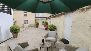 An outdoor patio area with wicker furniture, a table with a bottle and glasses, and a green umbrella. - Bluebell Cottage (Tenby)