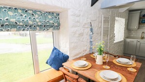 A dining area with a wooden table set for four, a blue cushion, and a view of a garden through a window with patterned curtains. - Bluebell Cottage (Tenby)