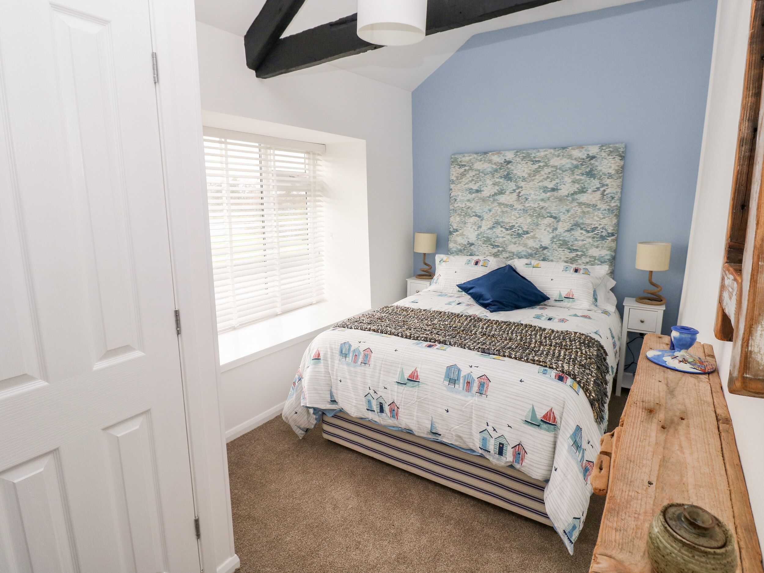A bedroom with a bed, a window with blinds, a headboard with a floral pattern, two bedside lamps, and a wooden nightstand with a blue cup.