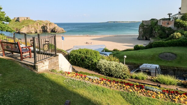 A coastal view with a beach, a grassy area with flowers, and a stone structure. - Bluebell Cottage (Tenby)